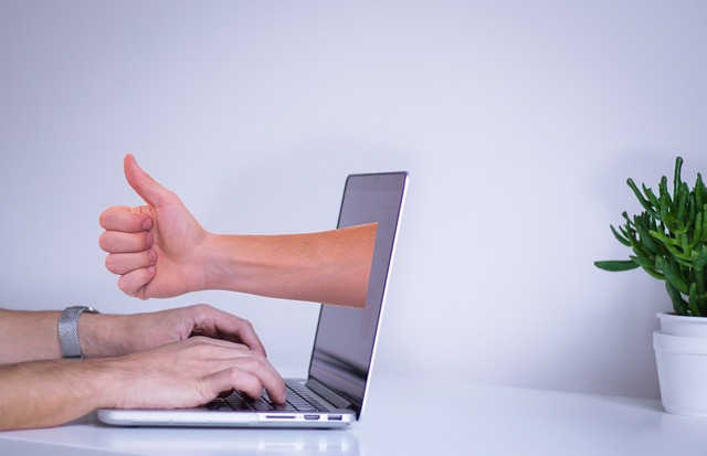 A hand giving a thumbs-up emerging from a laptop screen beside a person typing.