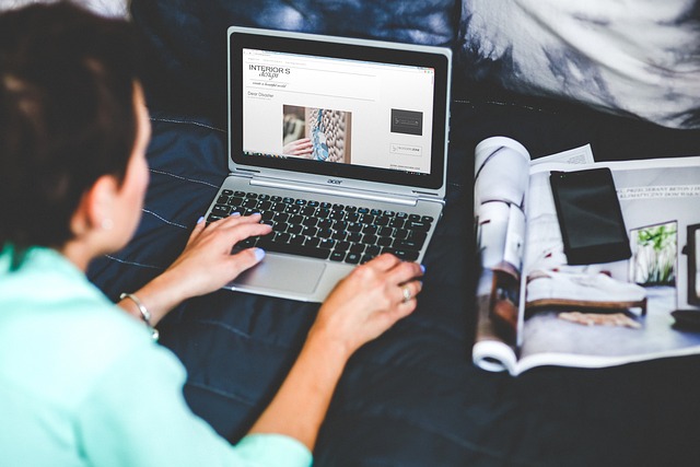 A person using a laptop to browse an interior design blog, with a magazine and smartphone placed on a nearby bed.